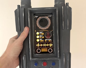 May include: A gray, rectangular prop with two cylindrical protrusions. It features a central screen displaying a digital interface with orange and yellow accents, and several buttons in blue, red, and white.