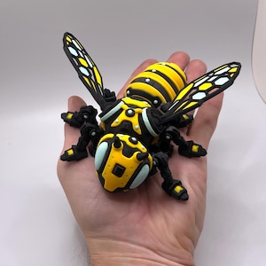 Op de afbeelding: Een gedetailleerd, geel en zwart insectenmodel met beweeglijke poten en vleugels. Het model heeft een gesegmenteerd lichaam, met gele en zwarte strepen. De vleugels hebben een zwart frame met gele en witte details. Het insect wordt in een hand gehouden.