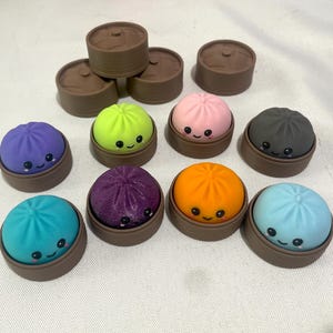 May include: A collection of small, round, colourful dumpling-shaped toys, each featuring a smiling face with black eyes and a small mouth. Each toy sits inside a brown, circular, basket-like base. The dumplings are purple, lime green, pink, dark grey, orange, light blue, and teal.