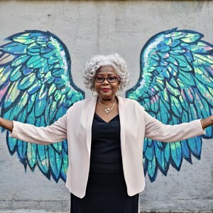 May include: An older woman with gray hair and glasses stands in front of a mural of angel wings. She wears a black dress, a light pink blazer, and gold jewelry. The wings are blue, green, and iridescent.