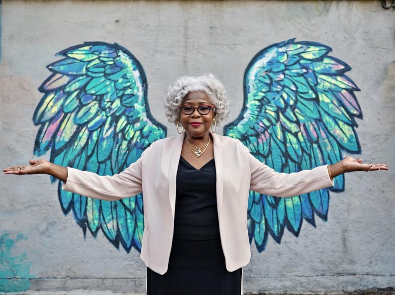 May include: An older woman with gray hair and glasses stands in front of a mural of angel wings. She wears a black dress, a light pink blazer, and gold jewelry. The wings are blue, green, and iridescent.