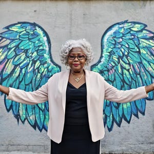May include: A person with curly grey hair wearing a light beige open-front blazer over a black top, accessorised with gold hoop earrings and a gold necklace, positioned in front of a mural featuring large, iridescent blue and green angel wings.