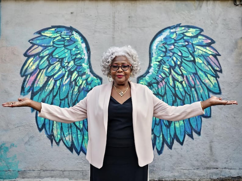 May include: A person with curly grey hair wearing a light beige open-front blazer over a black top, accessorised with gold hoop earrings and a gold necklace, positioned in front of a mural featuring large, iridescent blue and green angel wings.