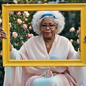 May include: A person with white hair, wearing a blue headband, glasses, and a gold necklace, holds a large rectangular yellow frame. The person is dressed in a light-coloured, flowing garment with soft pastel tones, positioned before a rose bush.