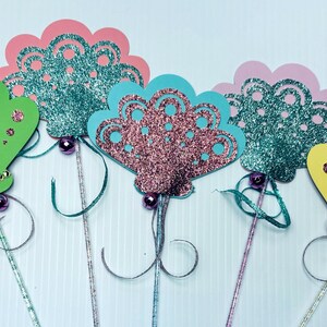 May include: A collection of colorful, glittery paper wands. The wands feature various shapes, including mittens and decorative fan designs, in shades of green, pink, blue, and yellow. Each wand has a decorative bead and ribbon.
