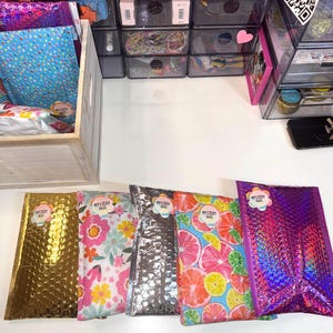 May include: A collection of colorful bubble mailers, including gold, floral, silver, citrus fruit, and iridescent purple. Each mailer has a "Mystery Bags" sticker. A wooden box and clear storage containers are in the background.