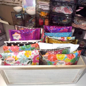 May include: A wooden box filled with colorful mystery bags. The bags have various patterns, including floral, polka dots, and fruit designs. Some bags are metallic and have the words "MYSTERY BAGS" printed on them.