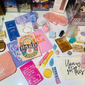 May include: An assortment of self-care items, including a pink eye mask, notebooks with motivational quotes, a headband, and a cosmetic bag with the text "You got this." Other items include a bracelet, bath salts, and skincare products.