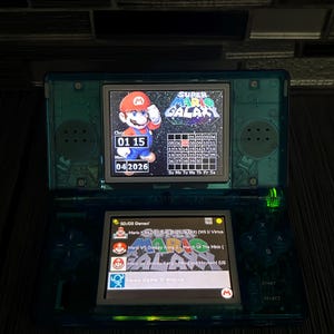 May include: A teal Nintendo DS Lite handheld game console, open and displaying game screens. The top screen shows a Super Mario Galaxy game calendar. The bottom screen shows a list of games. The date displayed is 01/15/2026.