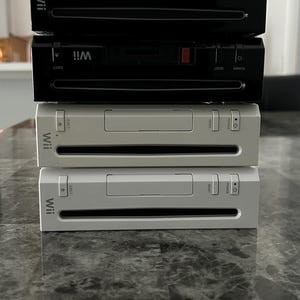 May include: Four Nintendo Wii consoles stacked on a grey surface. Two consoles are black, and two are white. Each console features the Wii logo, an eject button, a reset button, and a power button. The consoles are stacked on a marble-effect surface.