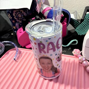 Puede incluir: Un vaso de acero inoxidable con tapa y pajita transparente. El vaso est&aacute; decorado con una foto de una mujer y el nombre "Grace" en lunares de colores. Se encuentra sobre una alfombrilla rosa acanalada.