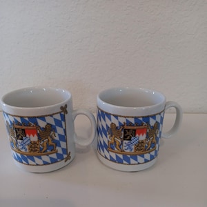 May include: Two white ceramic mugs with a blue and white diamond pattern and a gold border. Each mug features a coat of arms with lions and a shield. The mugs have a handle and are suitable for hot beverages.