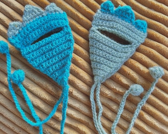 May include: Two handmade crochet bookmarks in the shape of a hand, one in turquoise and the other in light green. Each bookmark has a long, braided cord with a small ball at the end. The bookmarks are placed on a textured, tan surface.