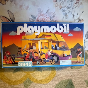 Puede incluir: Caja de Playmobil con una autocaravana amarilla y morada con figuras, bicicletas y una mesa. La caja tiene el logotipo de Playmobil y el número 3945. El fondo muestra una escena de atardecer.