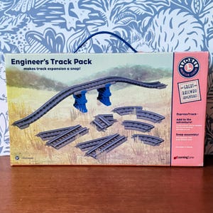 May include: A box set of Lionel Engineer's Track Pack, featuring gray train tracks, blue supports, and the text "Engineer's Track Pack" and "ExpressTrack". The box has a handle and a pink side panel with the words "Great Railway Adventures".