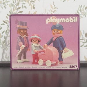 Könnte beinhalten: Eine Playmobil-Box mit einer vierköpfigen Familie. Die Figuren umfassen einen Mann mit Zylinder und Gehstock, eine Frau mit Handtasche, ein Kind mit Kinderwagen und ein weiteres Kind. Die Box ist lila mit dem Playmobil-Logo und der Nummer 5507.