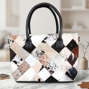 Puede incluir: Un bolso con un diseño de patchwork de cuadrados de piel de vaca en varios tonos de blanco, marrón, negro y beige. El bolso tiene dos asas de cuero negro unidas a la parte superior, una base rectangular y una silueta estructurada con pelo.