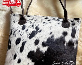 May include: A cowhide leather tote bag with a black and white spotted pattern. The bag has brown leather handles and trim. The text "Cowhide Leather Tote Bag" is written on the bottom right corner.