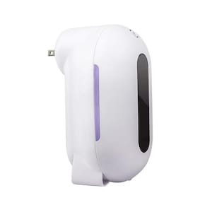 Puede incluir: Un ambientador blanco enchufable con un diseño redondeado. Cuenta con una ventana vertical negra y una luz púrpura visible a través de un panel translúcido. El dispositivo tiene una base pequeña y curvada y un enchufe en la parte superior izquierda.