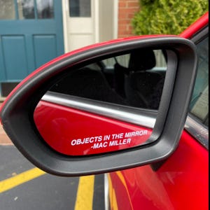 Op de afbeelding: Rode zijspiegel van een auto met een zwarte rand. De spiegel toont de tekst "OBJECTS IN THE MIRROR -MAC MILLER" in het wit. De spiegel reflecteert een gedeeltelijk zicht op het interieur van de auto en de omgeving.