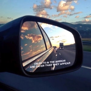 May include: A car's side mirror reflects a sunset with fluffy clouds and the text "OBJECTS IN THE MIRROR ARE GAYER THAN THEY APPEAR." The road ahead includes other vehicles and a truck.