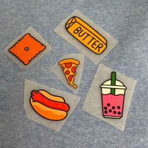 May include: A collection of iron-on patches featuring food illustrations. Includes a butter stick with the word "BUTTER", a pizza slice, a hot dog, a bubble tea, and a square orange cracker.