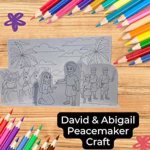May include: A gray paper craft featuring a biblical scene with the text "David & Abigail Peacemaker Craft." The scene depicts figures, donkeys, and palm trees. Colored pencils in various colors surround the craft on a wooden surface.