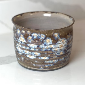 Puede incluir: Un cuenco de cerámica hecho a mano con un diseño texturizado y en capas. El cuenco presenta una base marrón oscuro con detalles en azul y blanco, creando un patrón único y orgánico. El interior es de un tono más claro y neutro.