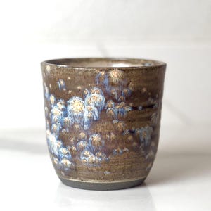 Puede incluir: Una taza de cerámica hecha a mano con un esmalte único. La taza presenta un color base marrón oscuro con patrones cristalinos azules y blancos. La taza tiene forma cilíndrica y un borde y base de color gris oscuro.