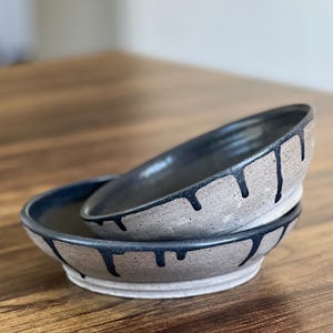 Pode incluir: Duas tigelas de cerâmica feitas à mão empilhadas sobre uma superfície de madeira. As tigelas são de cor bege claro com um interior azul escuro e um design de gotejamento ao redor da borda. As tigelas são provavelmente usadas para servir comida.