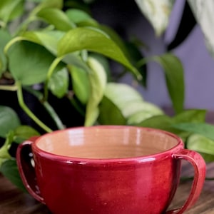 Puede incluir: Un cuenco de cerámica roja hecho a mano con dos asas. El interior del cuenco es de un color arcilla natural más claro. El cuenco está sobre una superficie de madera, con follaje verde en el fondo.