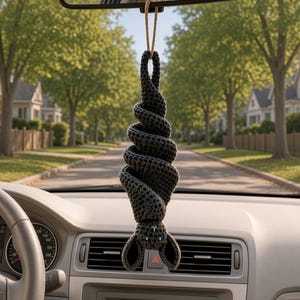 May include: A dark gray crocheted bat hanging upside down from a tan string. The bat features a spiral-shaped body, two pointed ears, and two small blue eyes. It is suspended from a rearview mirror inside a vehicle with a view of a suburban street.