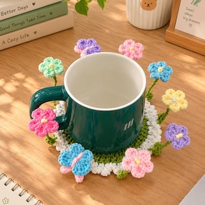May include: A dark green ceramic mug sits on a round, crocheted coaster featuring a white center, a green border, and various colorful crocheted flowers and butterflies attached to the edges in shades of pink, blue, yellow, and purple.