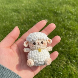 May include: A handmade, crocheted sheep figurine. The sheep is white with a fluffy texture, small beige ears, and a gold-coloured necklace with a star pendant. The sheep is sitting on a hand, with a green background.