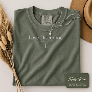 May include: A moss green short-sleeve t-shirt featuring the white text Love Discipline Proverbs 12:1 and a small cross icon. A silver star pendant necklace rests on the neckline. The shirt label reads Comfort Colors, 100% cotton, size S P CH.