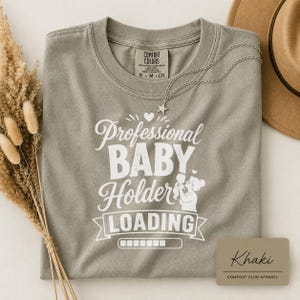 May include: A khaki-colored crew neck t-shirt featuring white screen-printed text that reads Professional BABY Holder LOADING with a graphic of a person holding a baby and a loading bar. The shirt has a Comfort Colors brand label at the neckline.