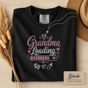 May include: A black crew neck t-shirt featuring the text Grandma Loading in pink and white lettering. The design includes a pink progress bar, small hearts, baby footprints, and a baby bottle graphic. The shirt has a Comfort Colors brand label.