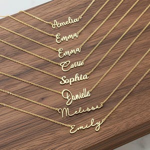 Könnte beinhalten: Goldfarbene Namensketten auf einer Holzoberfläche. Jede Halskette zeigt einen anderen Namen in Schreibschrift: Amelia, Emma, Carrie, Sophia, Danielle, Melissa und Emily. Die Halsketten sind zart und elegant.