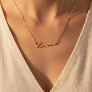 Puede incluir: Un collar dorado con el nombre "Lauren" en escritura cursiva. El collar se lleva alrededor del cuello, con la cadena y el colgante del nombre visibles sobre una parte superior de color claro.
