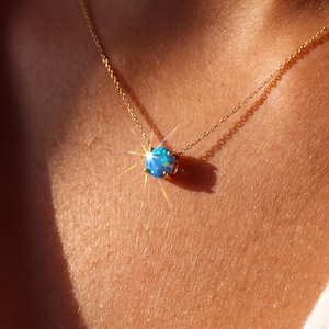 May include: A delicate gold chain necklace featuring a single round, iridescent blue opal pendant. The gemstone is set in a simple four-prong setting, catching the light with a bright, star-like sparkle against the warm, sunlit skin tone.