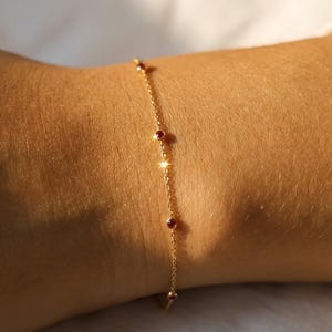 Op de afbeelding: Een fijne gouden armband met kleine, ronde, rode edelstenen gelijkmatig verdeeld over een fijne ketting. De armband wordt getoond op een arm, met een wit overhemd op de achtergrond. Een eenvoudig en elegant sieraad.
