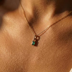 May include: A gold necklace with a small pendant featuring a rectangular emerald-green gemstone and small clear stones. The necklace is delicate and sits on a person's skin, with the chain visible.