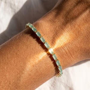 May include: A gold bracelet with rectangular, light blue gemstones. The bracelet is worn on a person's arm, with the gemstones arranged in a linear pattern. The bracelet is a delicate piece of jewelry.