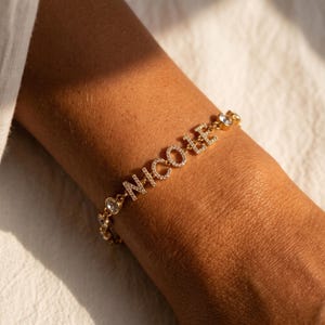 Könnte beinhalten: Goldfarbenes Armband mit dem Namen "NICOLE" in funkelnden, klaren Kristallen. Das Armband hat zusätzliche Akzente aus klaren Kristallen und wird am Handgelenk getragen. Ein stilvolles Schmuckstück.