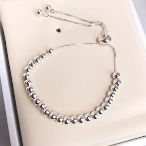 May include: A silver bracelet featuring a series of small, round beads. The bracelet has a sliding clasp for adjustable sizing and is displayed on a cream-colored surface. The design is simple and elegant.