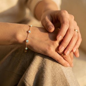 Puede incluir: Una delicada pulsera de cadena dorada con cuatro piedras de ópalo redondas, engastadas en bisel y espaciadas uniformemente, que muestran destellos iridiscentes azules y verdes. La pulsera presenta una fina cadena de eslabones dorados.