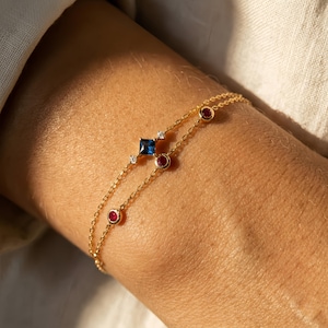Może przedstawiać: Delikatna złota bransoletka z dwoma sznurkami, ozdobiona kwadratowym niebieskim kamieniem szlachetnym, małymi białymi kamieniami i okrągłymi czerwonymi kamieniami. Bransoletka jest noszona na nadgarstku, prezentując wzór i kontrast kolorów biżuterii.