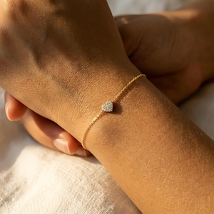 May include: A delicate gold bracelet with a small, heart-shaped charm. The charm is encrusted with small, sparkling stones. The bracelet is worn on a wrist, and the background is softly blurred.