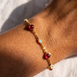 May include: A gold chain bracelet featuring alternating round deep red gemstones and small oval iridescent white opals. The delicate metal links connect each stone in a repeating pattern, creating a shimmering piece of jewelry with a polished finish.