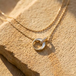 Peut inclure: Un collier doré à double chaîne. Le pendentif est composé d'un élément blanc en forme de croissant et d'un anneau orné de petites pierres transparentes. Le collier est présenté sur une surface texturée de couleur beige.
